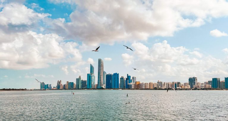 Stunning view of Abu Dhabi skyline with birds flying above the sea under a cloudy sky.