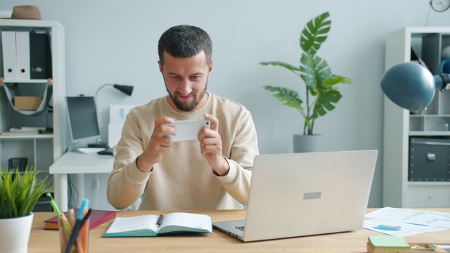 Man using a smartphone at an office desk with a laptop